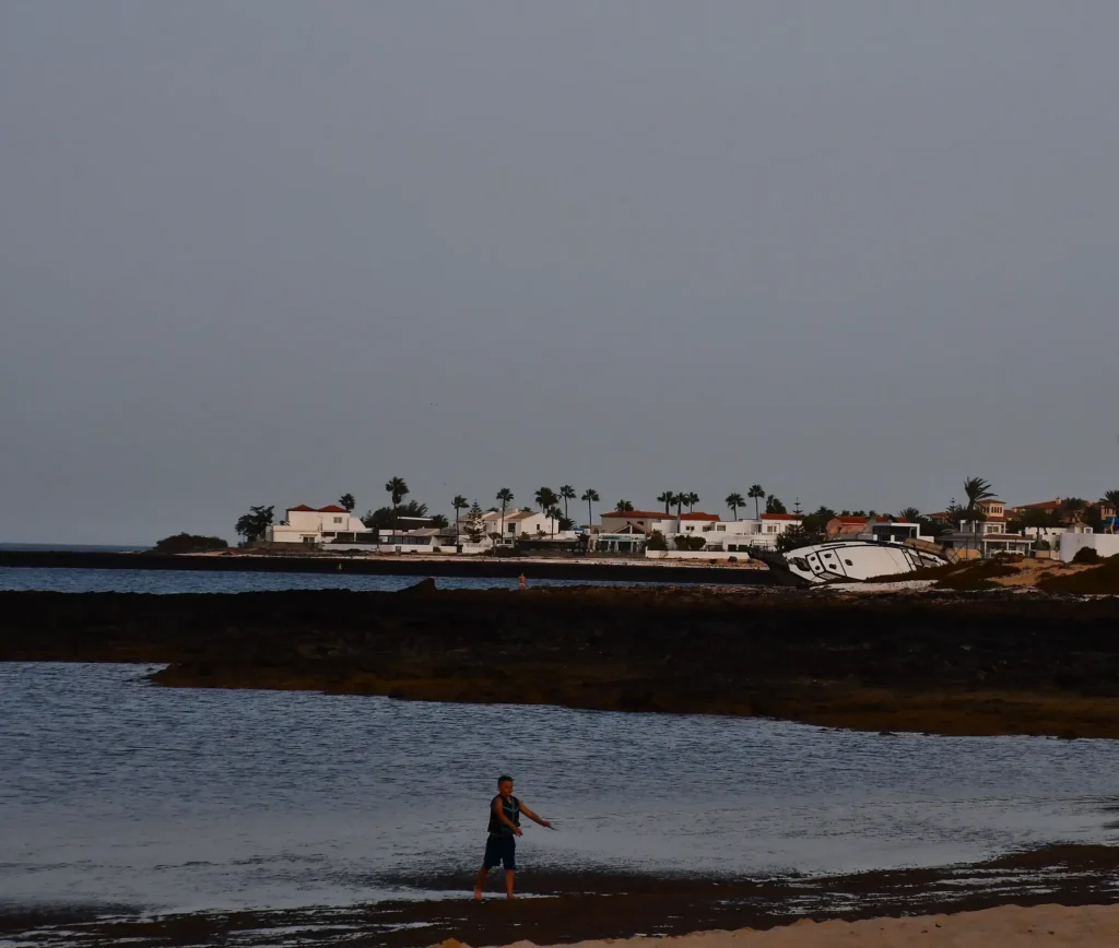 Un jeune garçon sort de l'eau en courant avec des maisons en arrière plan et au second plan nous pouvons apercevoir un bateau qui se repose sur sa coque