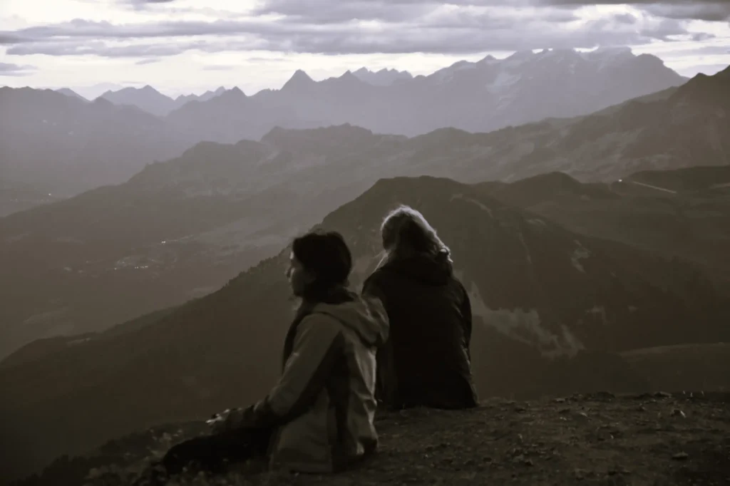 deux personnes assisent au sommet d'une montagne devant un paysage en attendant le lever du soleil