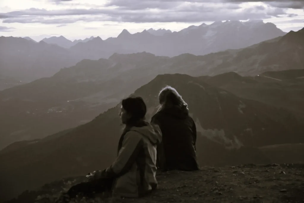 deux personnes assisent au sommet d'une montagne devant un paysage en attendant le lever du soleil