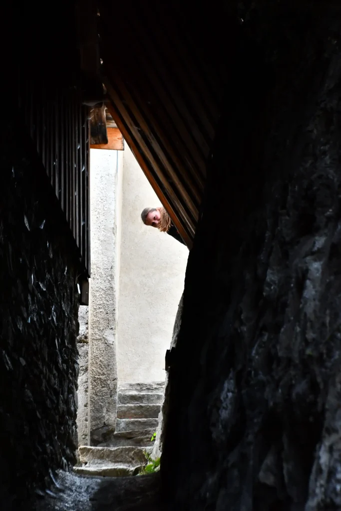 Un couloir extérieur en pierre avec des escaliers au bout et une tête d'une jeune femme qui apparait au bout