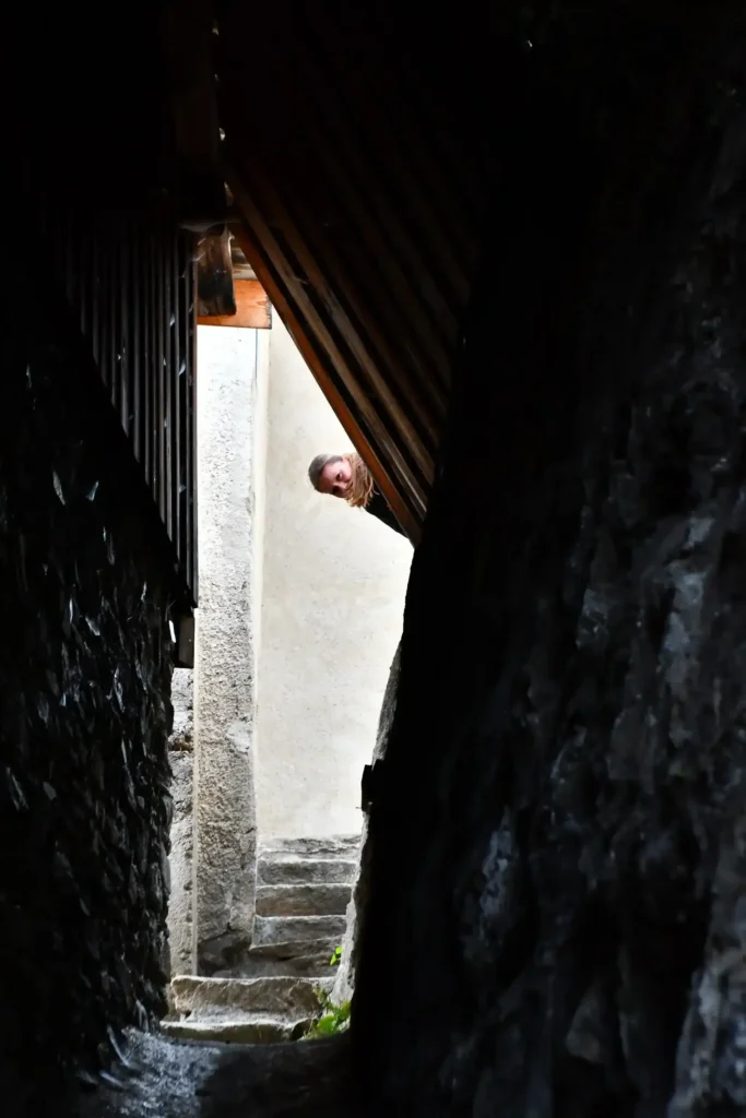 Un couloir extérieur en pierre avec des escaliers au bout et une tête d'une jeune femme qui apparait au bout
