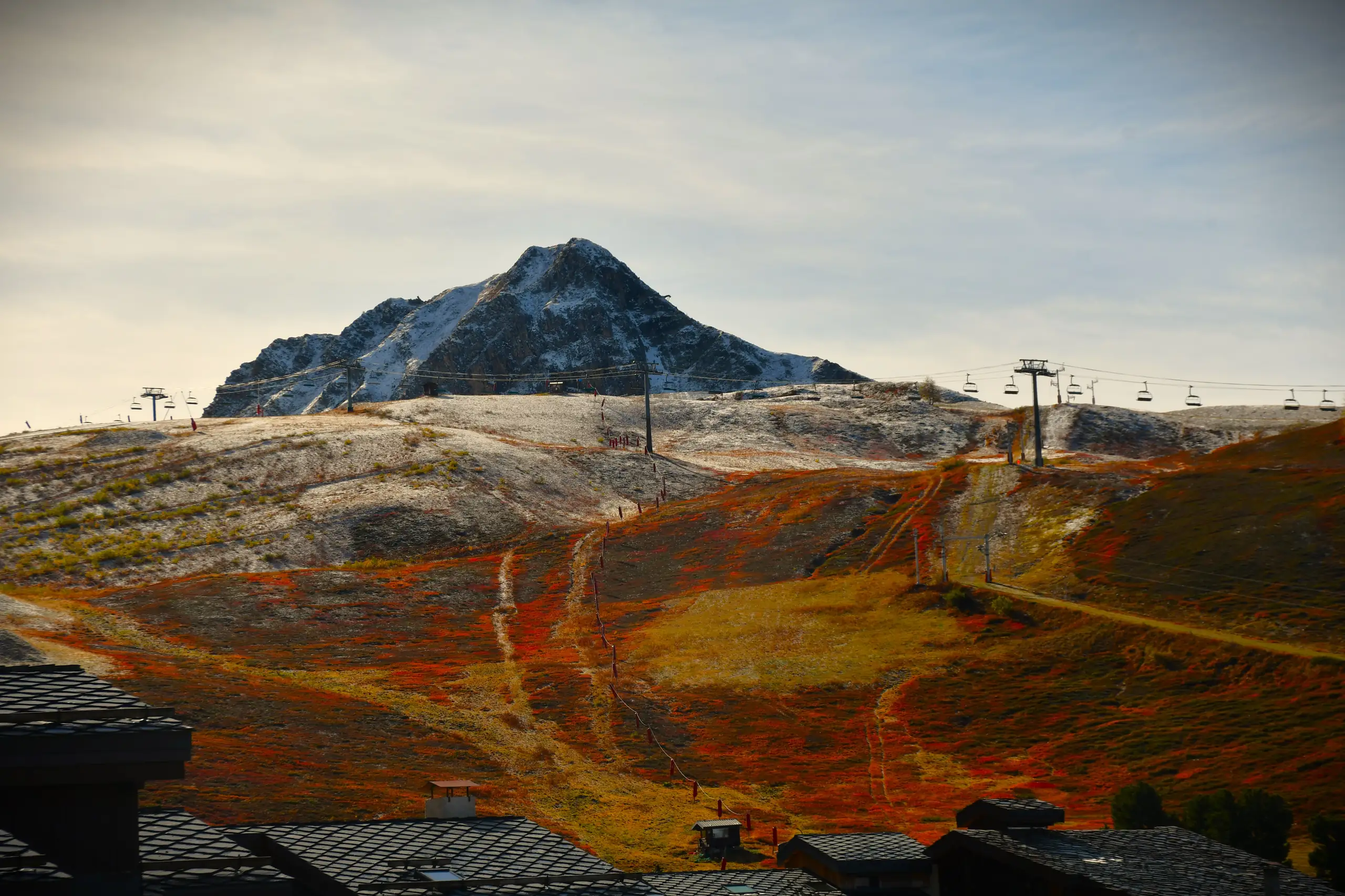 l'automne à la montagne, paysage entre sol et sommet saupoudrés de neige et remontée mécanique ainsi que des toits de chalet au premier plan