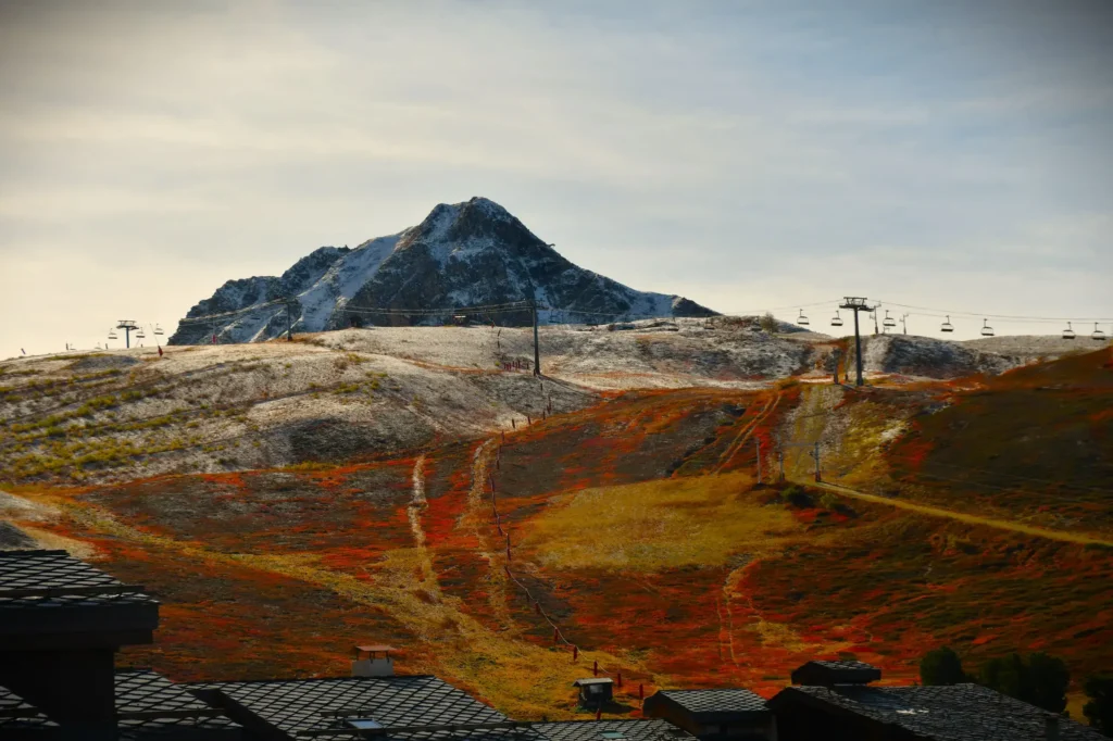 l'automne à la montagne, paysage entre sol et sommet saupoudrés de neige et remontée mécanique ainsi que des toits de chalet au premier plan