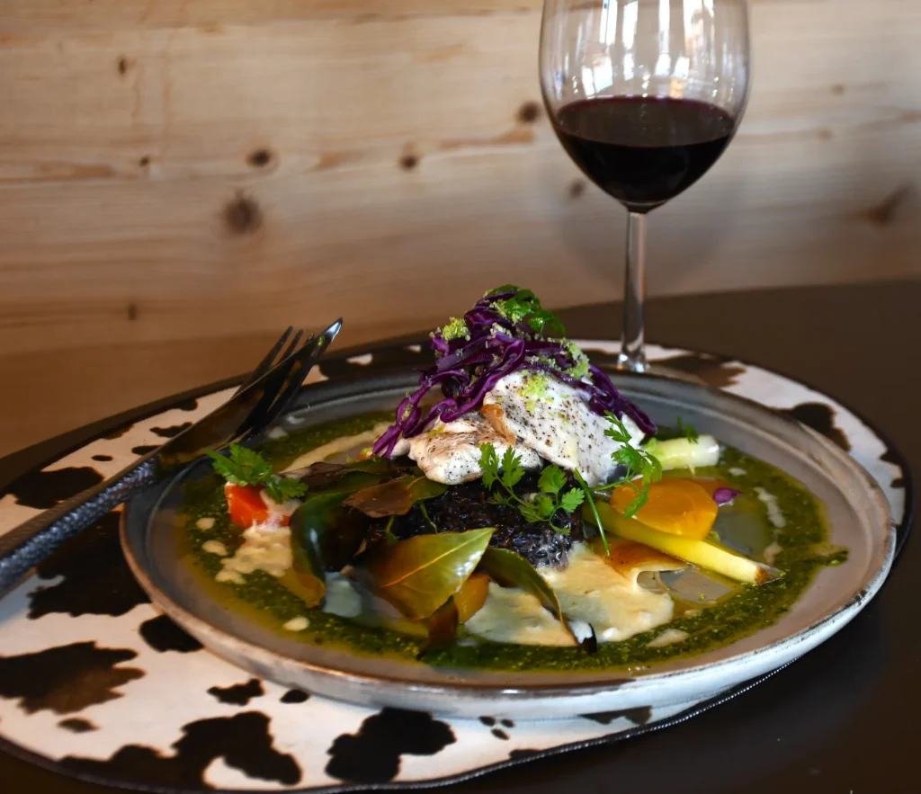 plat de restaurant : omble chevalier accompagné de son riz noir et de ses légumes avec un verre de vin rouge
